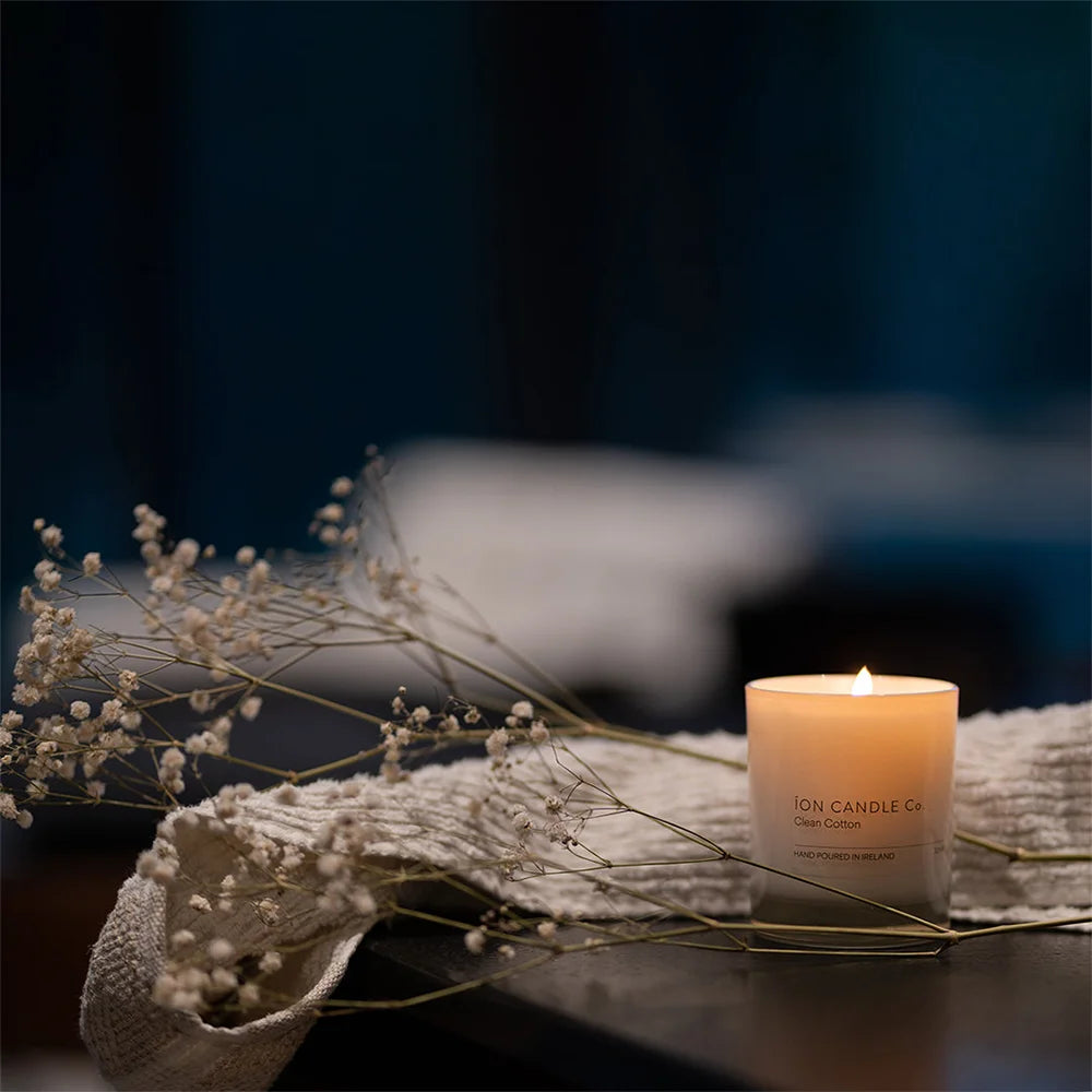Ion Candle Co. - Clean Cotton Luxury Scented Candle placed on a wooden desk next to some wispy flowers. It looks very nice and elegant. The background is a blurry navy blue