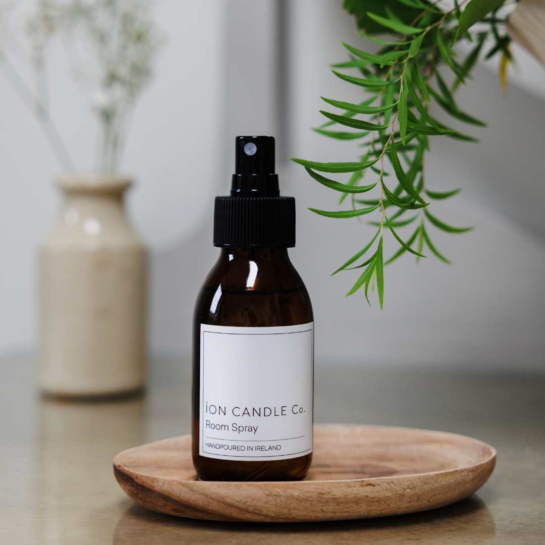 Ion Candle Co. - Luxury Scented Room Spray 100ml bottle against a white background with green foliage and vase. The bottle is brown and transparent with a white label and black text.