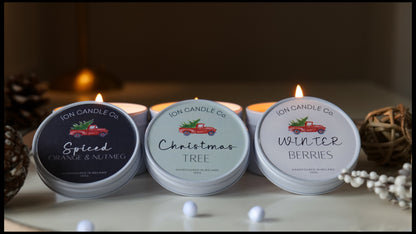 Three round metal candles with lids open, arranged on a tabletop in a warm indoor setting. The candles are labeled Spiced Orange & Nutmeg, Christmas Tree, and Winter Berries by 10H Candle Co.