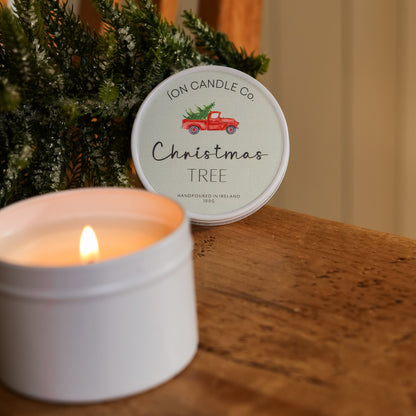 A white tin candle with a lit flame sits on a wooden table next to its lid, which features a round label reading 'ION CANDLE Co Christmas TREE' with a small red truck graphic. Green pine branches decorate the background.