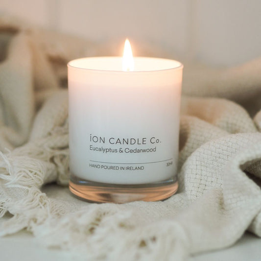 Ion Candle Co. -  luxury scented eucalyptus & cedarwood candle placed on a white cotton throw. The candle is lit and it looks very cosy.