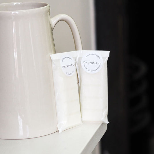 Ion Candle Co. - luxury scented Eucalyptus & cedarwood wax melts in their white paper packaging placed on the edge of a shelf next to a white ceramic pottery jug