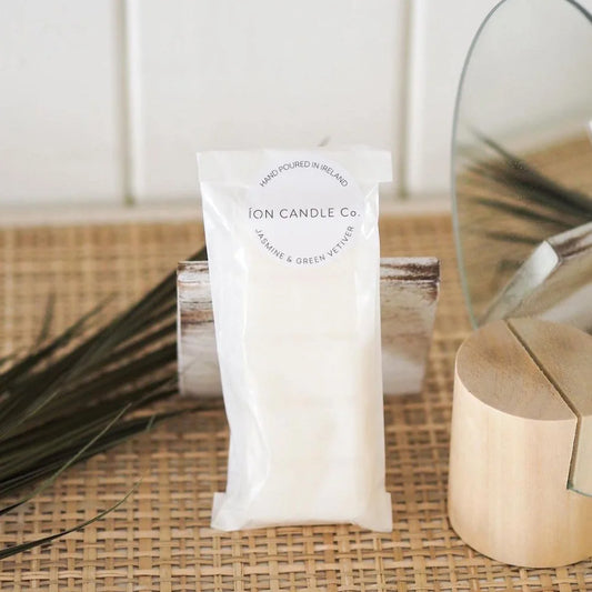 Ion Candle Co. - Luxury scented jasmine and green vetiver wax melts, in their white paper packaging placed a wicker straw shelf next to various wooden items with some grasses in the foreground