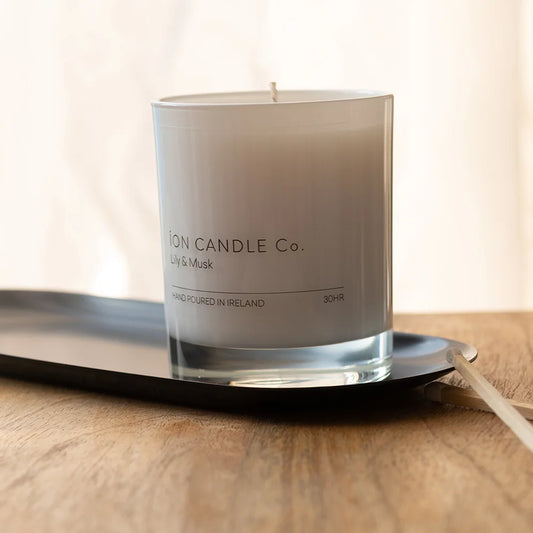 Ion Candle Co. - Lily and Musk Luxury Scented Candle, placed on a black oval shaped candle tray, with a match lying next to it. 