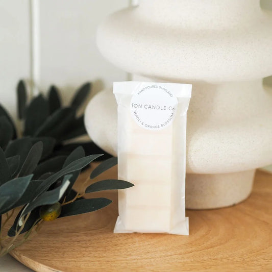 Ion Candle Co. - Luxury scented Neroli & orange blossom wax melts, in their white paper packaging leaning up against a white oddly shaped vase with some foliage to the left of shot