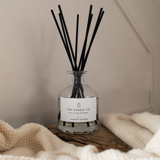 Reed diffuser with black sticks in a clear bottle on a wooden surface with a soft blanket in the background.