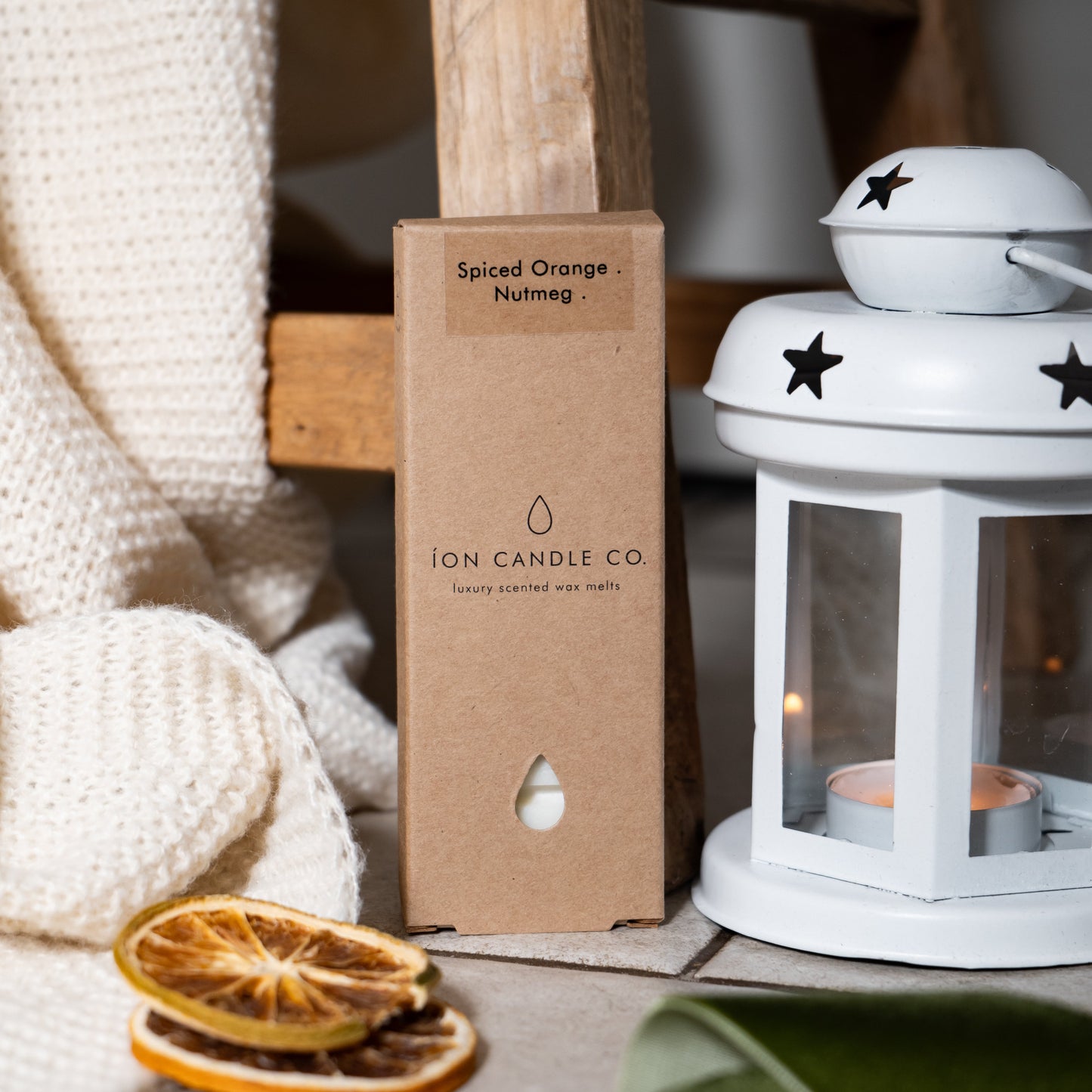 Spiced Orange Natoug packaging by Ion Candle Co with a white lantern and dried oranges on a wooden surface.