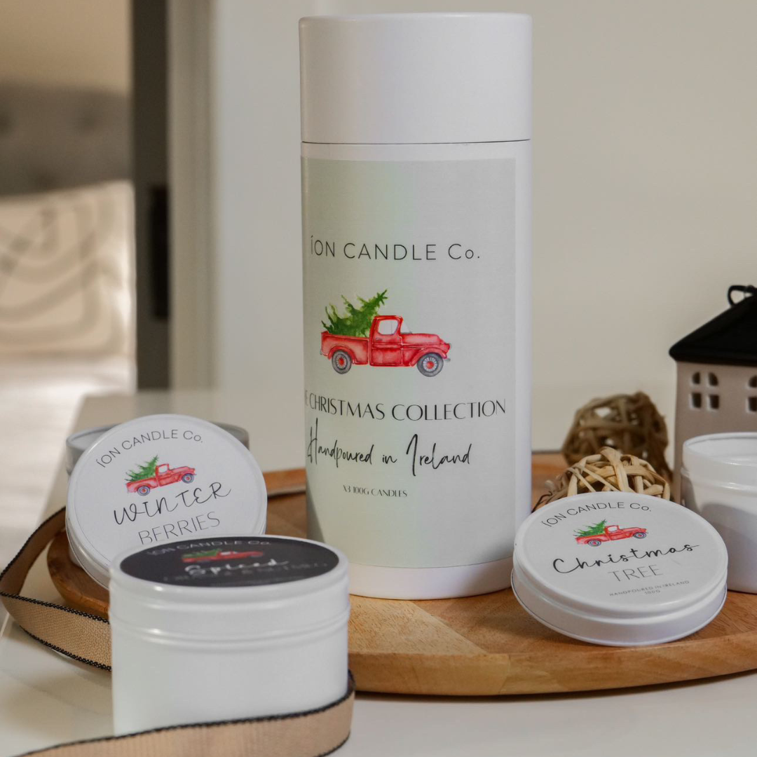 A set of three travel-sized candles from the Christmas Collection by Ion Candle Co., displayed in a festive gift tube with holiday-themed decorations around it.